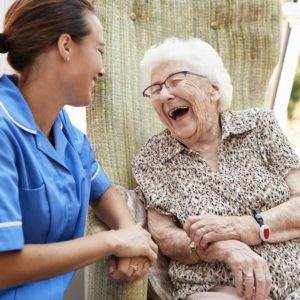 Senior Woman Sitting In Chair And Laughing With Nurse In Retirement Home